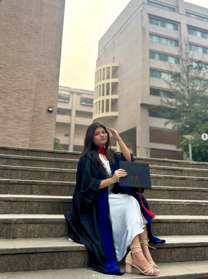 Mubashwira in a graduation gown, sitting on campus steps holding a mortarboard